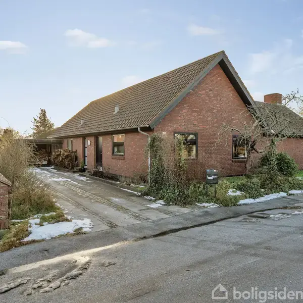 Hus i mursten med skråt tag står ved en vej. Snepletter dækker den omgivende indkørsel og buske. En bil på indkørslen og sparsom vegetationsvækst omkring.