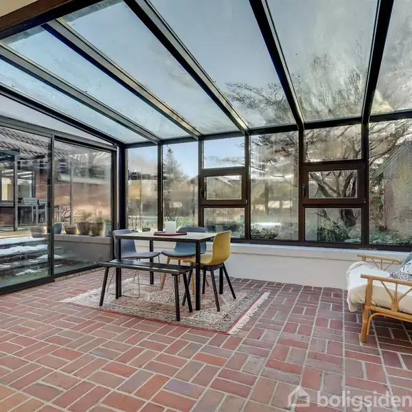 Et solbeskinnet udestue med glasvægge indeholder et bord med to stole, en gul og en blå. En rattan-stol står ved siden af, og klinkegulvet understreger den hyggelige atmosfære.