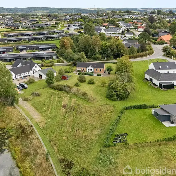Nabolag med moderne huse omgivet af grønne arealer og en lille sø. Huse ligger langs veje med græsplæner, træer og stisystemer i et naturskønt, bebygget område.