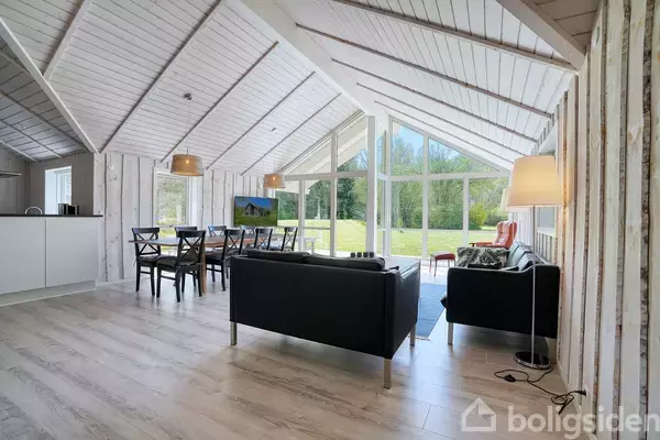 Sort sofa i moderne stue vender mod store glasvægge, indrammet af hvide træpaneler og loft. Til venstre er et spisebord med stole. Udenfor er der en grøn have.