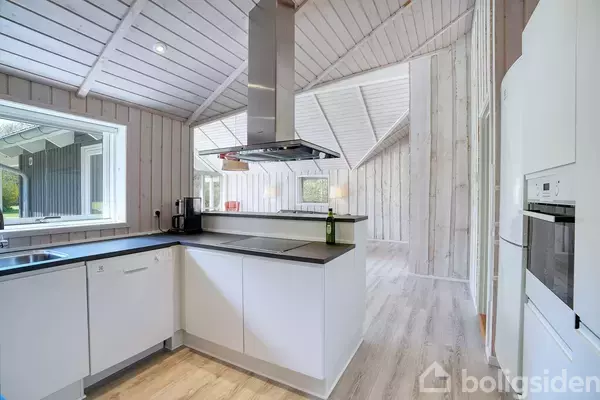 Hvidt køkken med vinkelformet bordplade, integreret kogeplade og emhætte. En vinflaske står på bordet. Store vinduer og lyst træpanel på vægge og loft i et åbent rum.