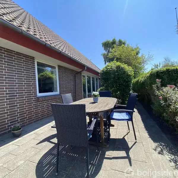 Et rundt bord med mørke stole står på en terrasse ved siden af en murstensbygning. Sollys skinner, og omgivelserne er præget af buske og træer.