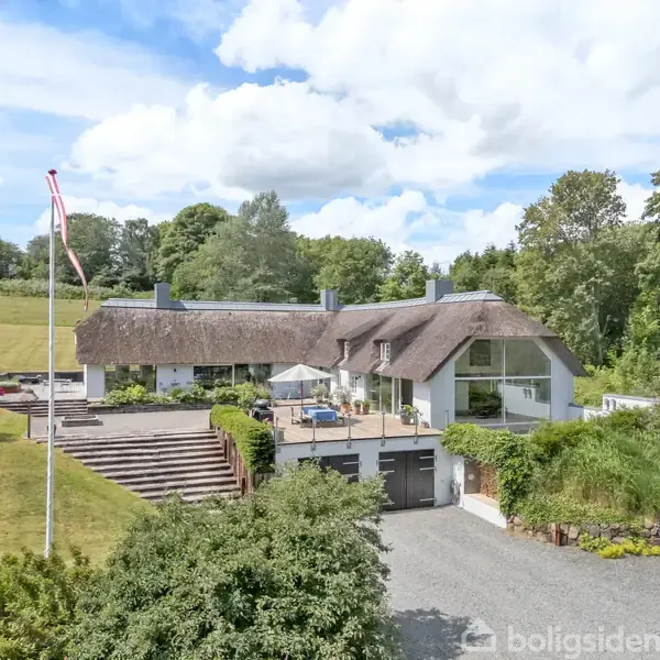 Stråtækt hus omgivet af grønne træer og en grusvej. Huset har et stort glasparti og en terrasse med parasol. En flagstang med flag står i forhaven.
