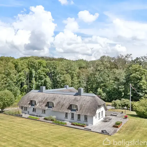 Stråtækt hus med hvid facade ligger i en stor, velplejet have. Baggrunden domineres af en tæt skov, og himlen er delvist skyet.