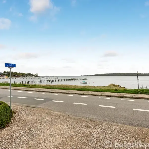 En vej med vejskiltet "7030" strækker sig langs en kystlinje med udsigt over vandet. I baggrunden ses en lang bådebro og spredte skyer på en blå himmel.