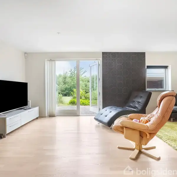 Stue med fladskærms-TV på skænk til venstre, lænestol og sort sofa i midten. Store vinduer og dør i baggrunden åbner ud til en have. Grønne tæpper og vægge.