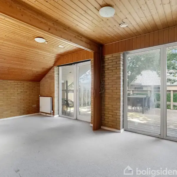 Et tomt rum med skrå træloft og lyse vægge, hvor sollys strømmer ind gennem store glasdøre, der åbner til en terrasse med udsigt til et hus og træer udenfor.