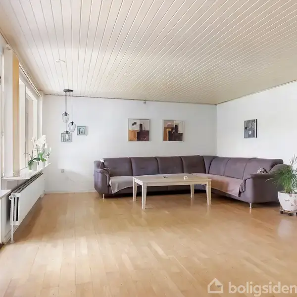 En stor stue med en L-formet grå sofa, der står langs hjørnevæggen. Et træbord er placeret foran sofaen. Rummet har lyse træpaneler og store vinduer med udsigt til naturen.