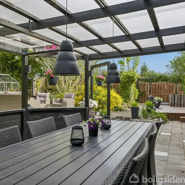 En overdækket terrasse med et langt spisebord og stole. Hængende lamper over bordet. I baggrunden ses en have med blomster, siddeområde og grill på fliser.