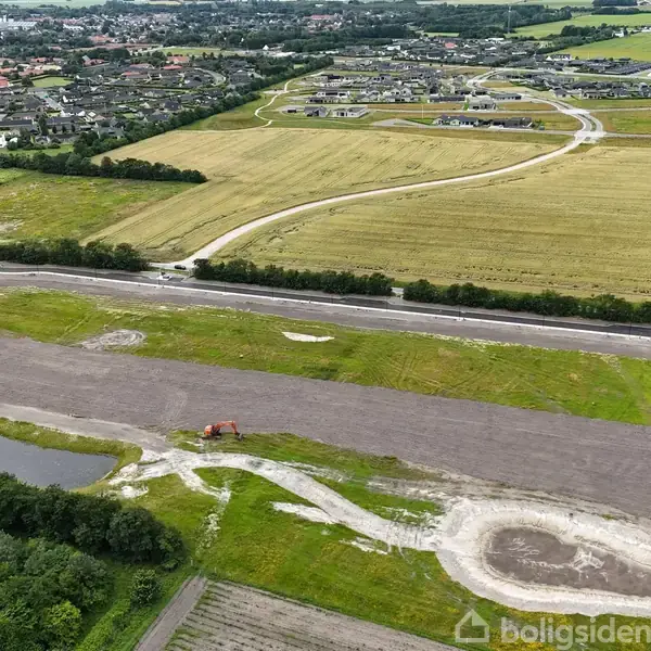 Gravemaskine arbejder på en delvist asfalteret byggeplads omgivet af marker og boligområder. Fjernliggende byområde og grønne marker ses i baggrunden.