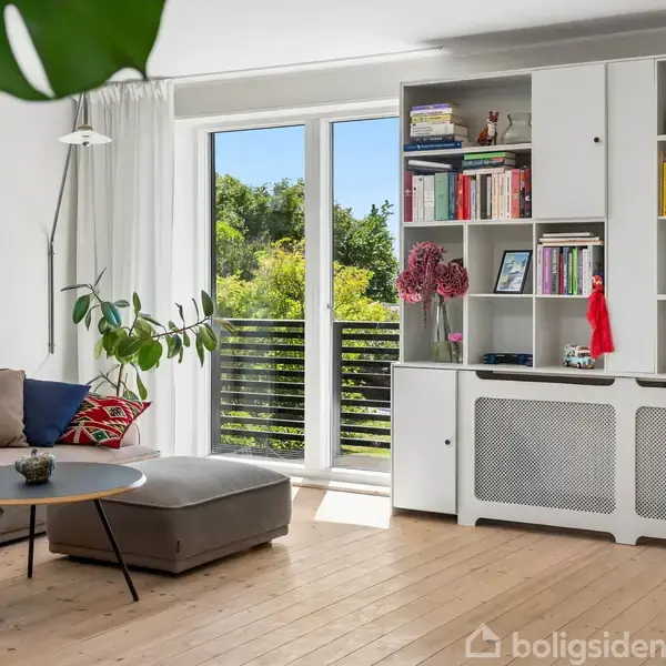 Stue med sofa og sofabord ved siden af en stor, grøn plante. Reol med bøger og dekorationer. Vindue med udsigt til frodige træer udenfor. Lys og moderne indretning.