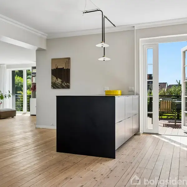 En moderne stue med trægulv og sorte køkkenskabe står centralt. En dør til højre er åben mod en terrasse med udsigt til grønne omgivelser. En sofa ses i baggrunden.