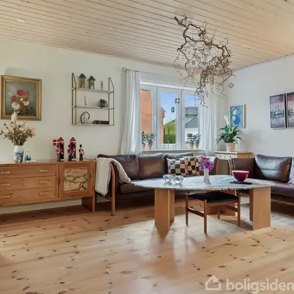 Stue med sort lædersofa i hjørne, firkantet træbord i midten, trækommode med lamper til venstre, store vinduer med hvidt gardin, flere dekorative elementer på vægge.