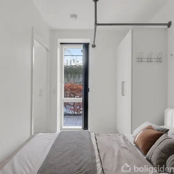 Soveværelse med en seng dækket af puder og et tæppe. Væggene er hvide, der er et klædeskab og en åben dør fører til en terrasse med udsigt til buske.