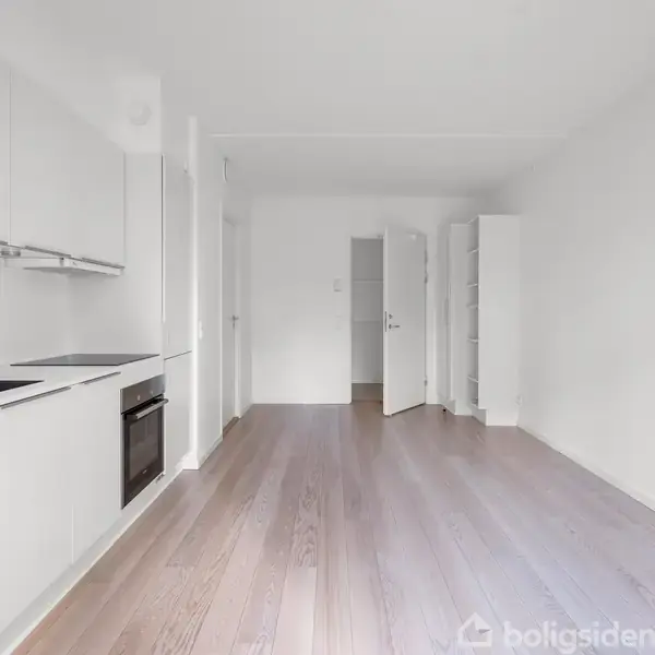 Et lyst, minimalistisk køkken med hvidt skabsarrangement og trælaminatgulv. Køkkenet har en integreret ovn og kogeplade. En åben dør fører til et tilstødende rum.