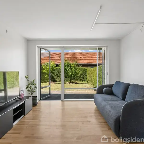 En stue med en grå sofa står til højre. Et fladskærms-tv er placeret på et lavt tv-bord til venstre. Glasdøre åbner ud til en have med hække.
