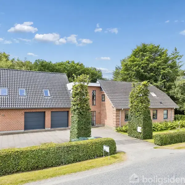 To rødkalkede, moderne murstenshuse med sorte tegltage står side om side omgivet af en frodig have og træer. Foran findes en flisebelagt indkørsel og veltrimmede hække.