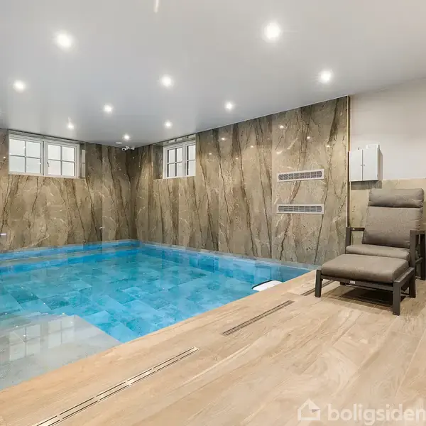 Indendørs swimmingpool med klart blåt vand, omkranset af naturfarvede marmorvægge. To polstrede stole står på et trægulv ved siden af poolen. Overheadbelysning oplyser rummet.