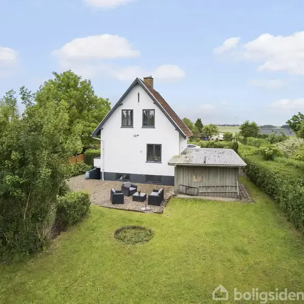 Et hvidt hus med spidst tag står i en grøn have. Huset har en terrasse med havemøbler. Omgivet af buske og træer, set under en blå himmel med få skyer.
