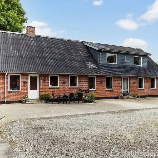 Et rødt murstenshus med sort tag står omkranset af grusvej og grøn vegetation. Foran huset er der sorte havemøbler opstillet på flisebelægning. Himmel er blå med lette skyer.