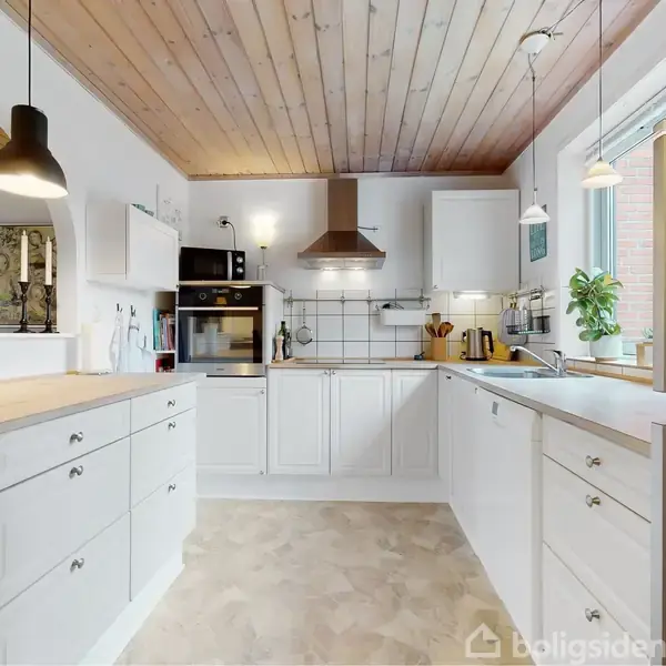Et moderne køkken med hvide skabe og træloft. Rummet er fyldt med naturligt lys fra et stort vindue, og der hænger lamper over arbejdsbordet. Der er ingen tekst.