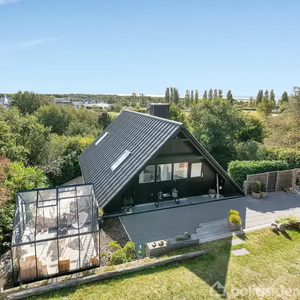 A-formet hus står stille, omgivet af frodig have og en overdækket terrasse. I baggrunden ses skov og marker, der skaber en fredelig og naturskøn atmosfære.