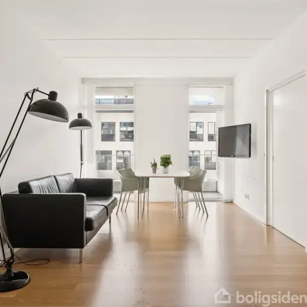 Sort sofa står ved siden af en stor gulvlampe og en spisebord med fire stole. Et fladskærms-tv hænger på væggen. Rummet er lyst med trægulve.