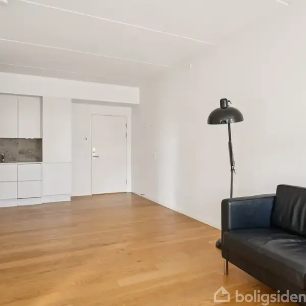 Sort sofa står stille i et lyst rum med trægulv. Til højre en buet standerlampe. Baggrunden viser et lille køkken og en hvid dør.