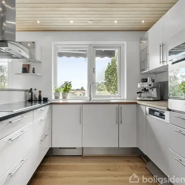 Lyst køkken med hvide skabe og sorte bordplader. Kaffemaskine står ved vindue med udsigt til have. Trægulve og indbyggede spotlights i loftet fuldender det moderne design.