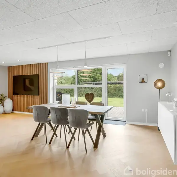 Spisebord med seks stole står i et lyst rum med store vinduer mod en grøn have. I baggrunden hænger et TV på en væg med dekorative vaser og malerier.