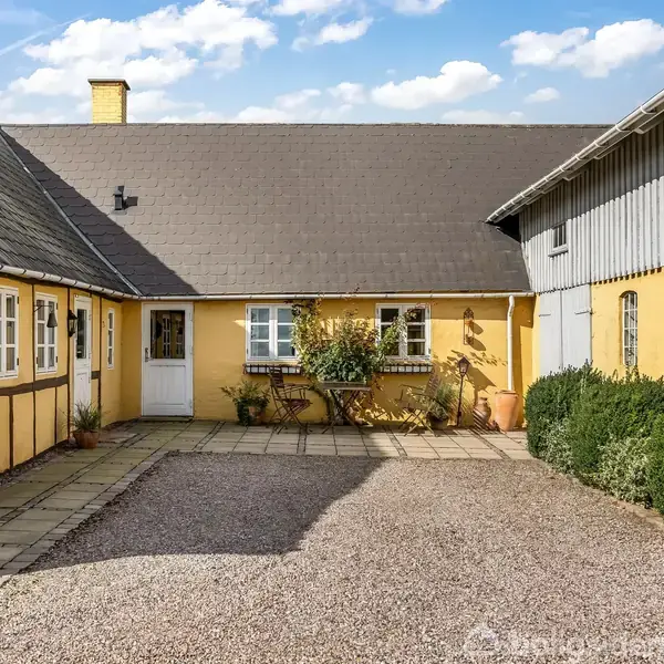 Hus i okkerfarve står stille i sollys, omgivet af gårdhave med brosten, blomster og buske. Bygningens tag er skifergrå, og der er flere vinduer med sprosser.