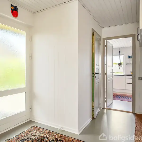 Entré med dør til køkkenet, hvor man kan se planter, en knagerække med tøj og en farverig tæppe på gulvet. En ugledekoration hænger over døren.