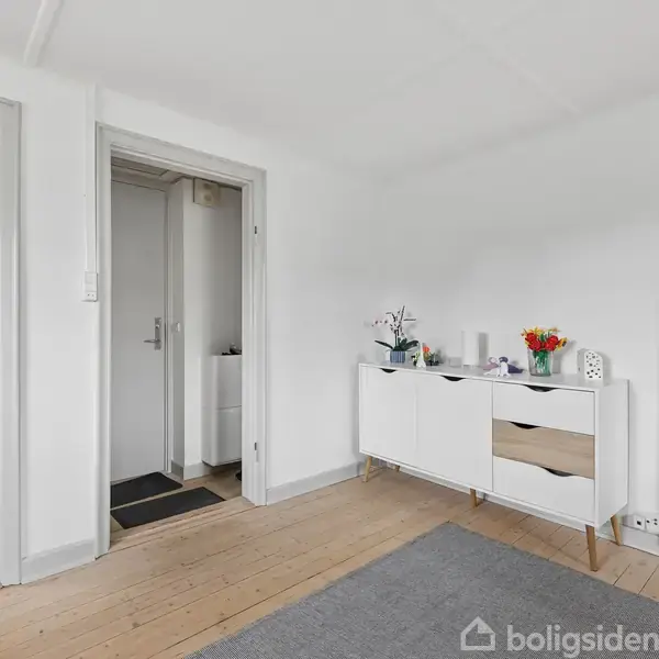 Hvid skænk står mod en væg med genstande som planter og bøger ovenpå. Skænkens skuffer er delvist åbne. I baggrunden ses en sofa i en lys, moderne stue.
