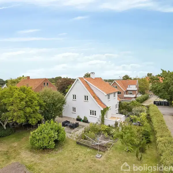 Hvidt hus med rødt tegltag står blandt grønne træer og buske. En flisebelagt terrasse ses ved siden af huset, og en smal vej løber til højre. Boligsiden-webstedet er vandmærket.