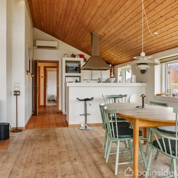 Pejs opvarmer stue med træloft, hvor et rundt bord og grønne stole står nær hvide køkkenelementer. Store vinduer lader lys ind, og gang leder til andre rum.