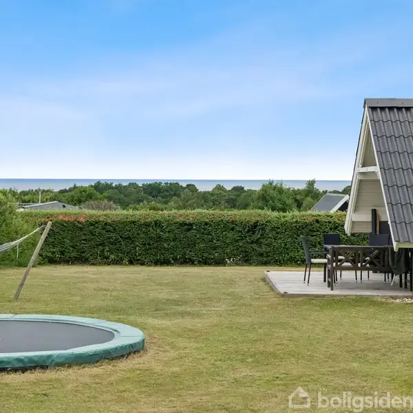 Trampolin ligger i en velplejet have, mens en badmintonbane strækker sig parallelt. I baggrunden står et hus med terrasse og borde, omgivet af hække og med udsigt over havet.