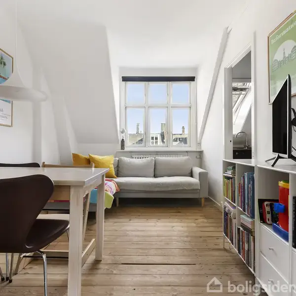 Sofa står foran et stort vindue med puder. Ved siden af er et spisebord med stole. Bøger og dekorationer på hylder skaber en hjemlig atmosfære.