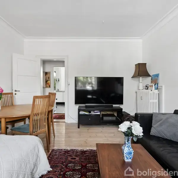 Stue med fladskærms-TV på skænk, sort sofa og sofabord med vase og blomster. Rundt spisebord med stole til venstre. Dekorativt tæppe på gulvet og kunst på væggen.