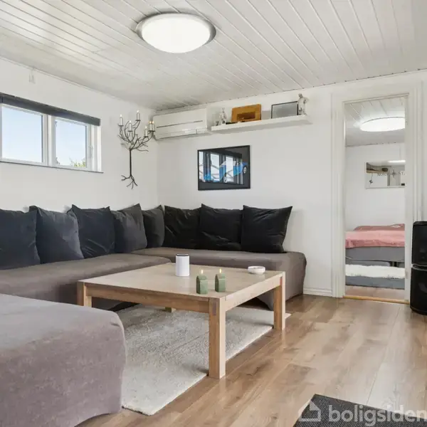 Stue med stor, grå hjørnesofa fyldt med puder. Et træbord står foran sofaen. Bag er et spejl og dør til et soveværelse. Vægge er hvide, gulv af træ.