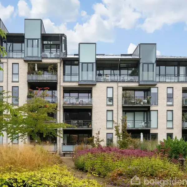 Lejlighedskompleks med flere etager, glasaltaner og moderne arkitektur. Beliggende i en grøn have fyldt med planter og blomster, med blå himmel og skyer i baggrunden.