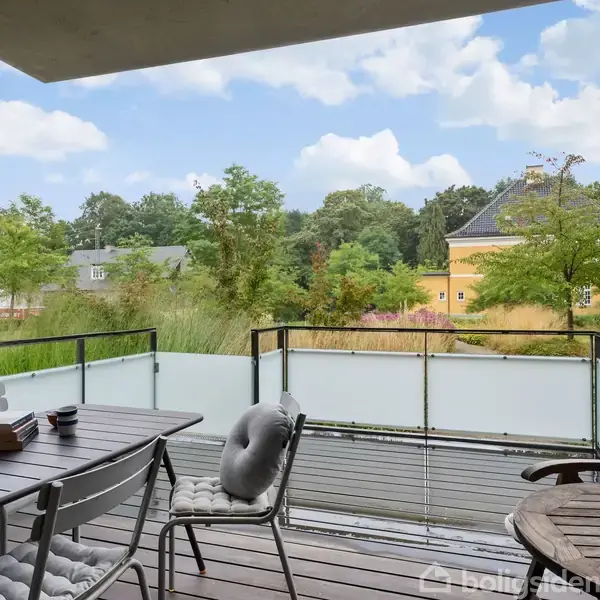 Terrasse med bord og stole med hynder, der står stille. Omkranset af glasrækværk, har terrassen udsigt til grønne træer og en gul bygning i baggrunden.
