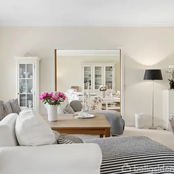 En lys stue med en hvid sofa, kaffe bord med blomster, og en stor fladskærms-TV. I baggrunden ses en skænk og et spisebord i et tilstødende rum.