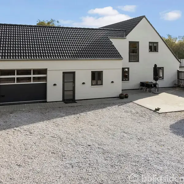 Et hvidt hus med sort tag står stille i en grusbelagt gårdsplads. Til venstre ses en integreret garageport. Haven i baggrunden er omgivet af grønne træer.