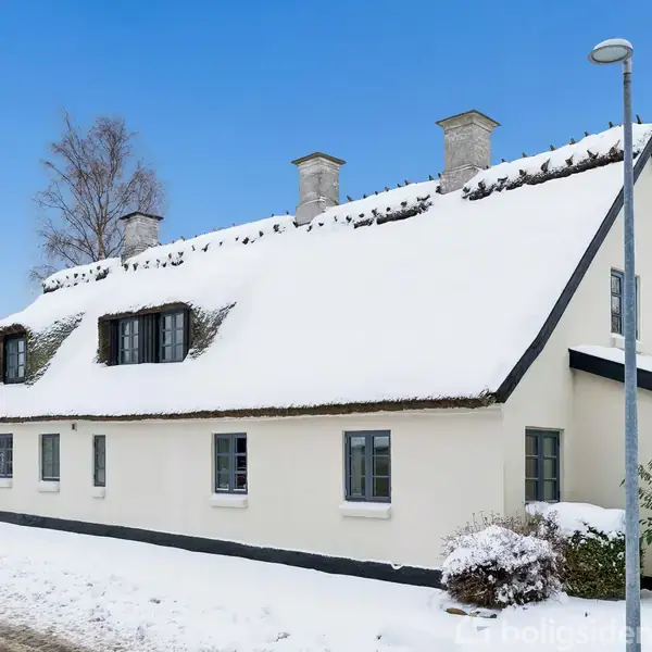 En hvidmalet, stråtækt hus står dækket af sne, med tre skorstene på taget. Det omgivende miljø er vinterligt med sne på jorden og en klar blå himmel.
