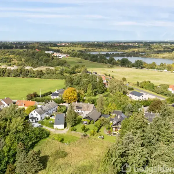 Landsbyhuse ligger tæt sammen omgivet af frodige skove og marker. I baggrunden ses en sø og åbne grønne områder, der strækker sig mod horisonten under en klar himmel.