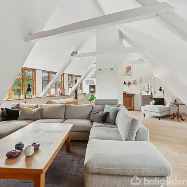 En stor, lysegrå sofa står i et rummeligt stueområde med skrå vægge. Et træbord er foran sofaen, og store vinduer giver udsigt til naturen udenfor.