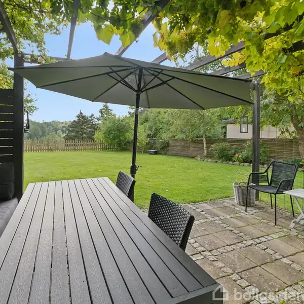 En sort spisebord med stole står under en stor parasol på en have-terrasse. Baggrunden viser en græsplæne omgivet af træer og buske.