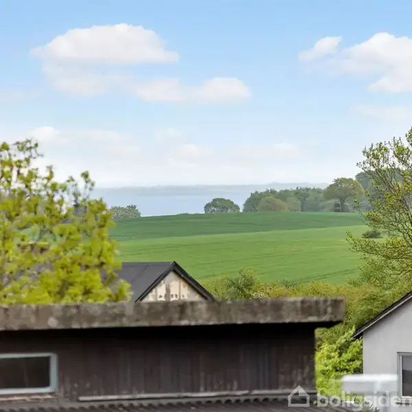 Bygninger i forgrunden står stille i et landskabsbillede, omgivet af grønne marker og træer med himmel og hav i baggrunden.