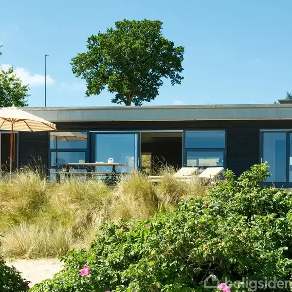 Et moderne sommerhus med store vinduer. Det står stille midt i en kystnær have med højt græs og buske, under en blå himmel. En parasol og havemøbler ses foran huset.