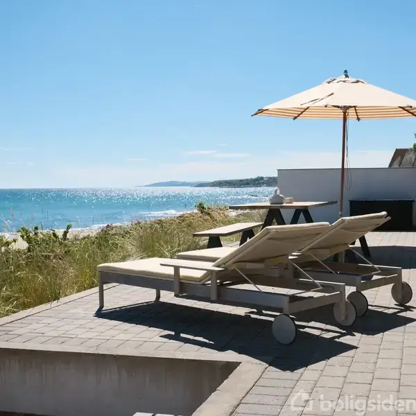 To liggestole står på en solrig terrasse med udsigt over havet. Et stort parasol skaber skygge, mens græsset langs klitterne bevæger sig let i vinden.